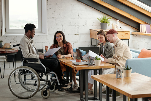 Two pairs of employees working in small groups by workplace while female economist pointing at document held by male colleague in wheelchair