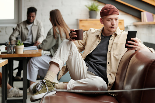 Young serious male white collar worker in casualwear texting in smartphone or looking through messages while sitting on couch