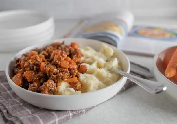 Simple home cooked meal with ground beef, carrots and mashed potatoes on a plate on kitchen counter background with a food magazine in the background.