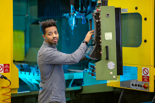A dark-skinned African-American man without safety equipment checks the settings and operation of a non-working automated computer-controlled metalworking machine, illuminated by a signal light, checking the quality of the drill head and drill bits on the machine. Portrait by the machine, copy paste, smiling worker,