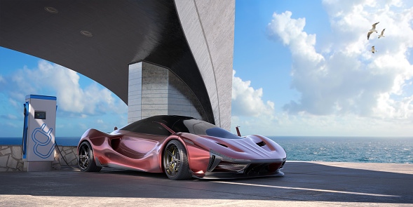A generic red electric sports car parked near the sea connected to an electric vehicle charging station, under an architectural arch bridge made from concrete and metal cladding. It is a bright day with clouds in the sky.