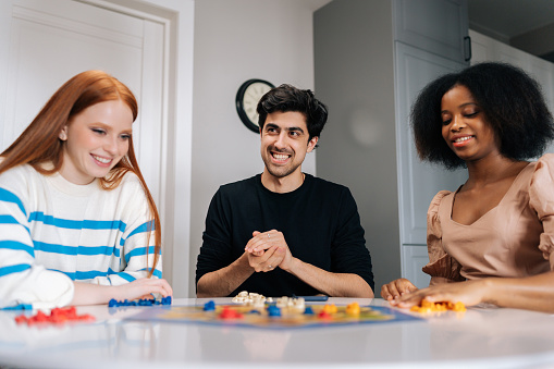 Portrait of cheerful young man throwing dice playing in board game with multiethnic friends, enjoying pastime leisure activity at home. Diverse happy people having fun and enjoying competition.
