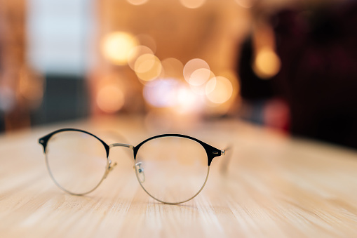 Selective focus of stylish glasses lying on wooden table on blurred bokeh background. Close-up of black classic eyeglasses lay on wooden desk with bokeh Christmas light.