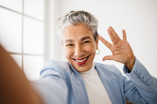 Senior business woman smiling and waving at the camera as she connects on a video call. Woman with graying hair using mobile technology to communicate with her clients and colleagues from her office.