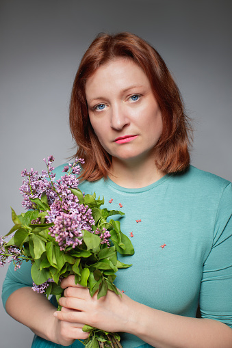 Portrait of mid adult woman with dyed red hair and blue eyes in turquoise top holding bunch of lilac flowers and looking at camera seriously, studio shot