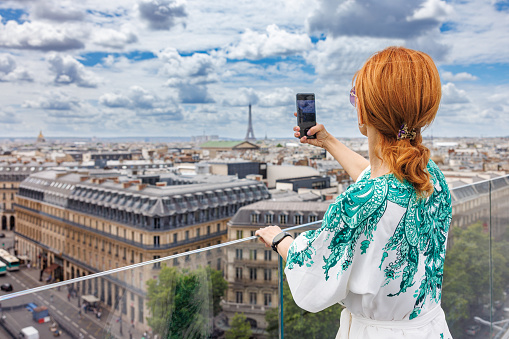 Woman standing on Galleries Lafayette building terrace by the glass fence and taking a photo of Paris cityscape under the cloudy sky with her mobile phone, technology usage lifestyle