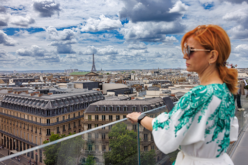 Elegant woman standing on a building terrace   of Lafayette gallery building by the glass fence and looking at Paris cityscape under the overcast, travel lifestyle