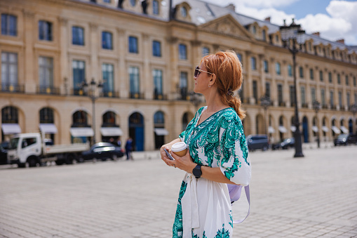 Elegant woman standing on a town square in Paris, looking and admiring architecture on a beautiful day, holding her mobile phone, travel lifestyle