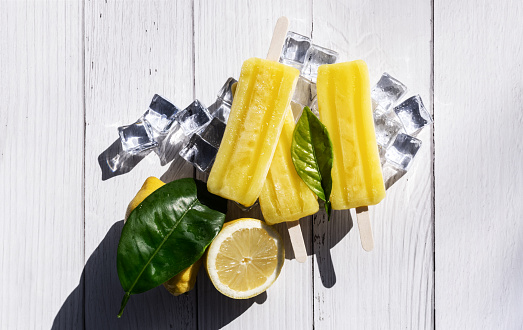 Yellow lemon popsicles or popsicles with wooden stick on ice cubes next to lemon leaves and slices on white wooden table with outdoor sunlight. View from above, overhead view. Cast hard shadows. Midday sun. Concept of cooling off, lemon popsicle, ice cream for the heat, hot summer day, rich ice cream, lemon flavor, ice cream.