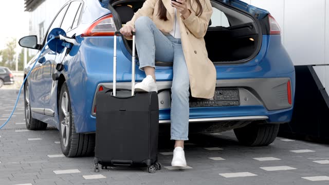 Young woman waiting charging automobile battery from small public station and using smartphone while charging automobile