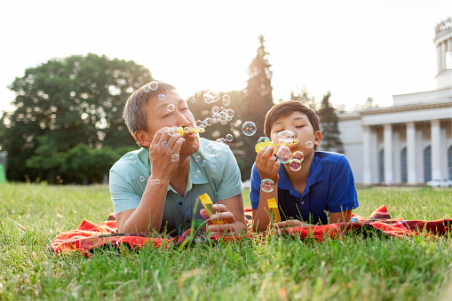 asian old father together with son lie on the grass in the park and blow soap bubbles and rejoice in summer, korean boy rest and play with dad outdoors, concept of parental love and care