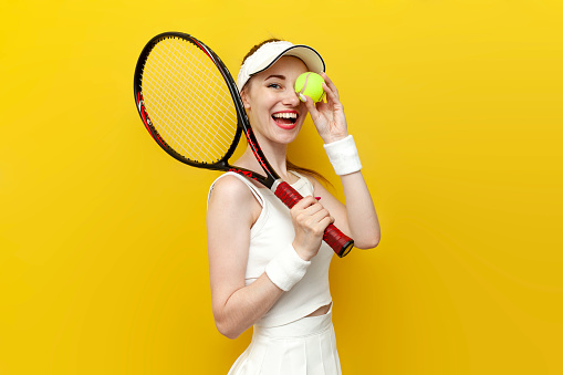 girl tennis player in sportswear holding tennis racket and ball on yellow isolated background and smiling, portrait of female tennis coach in white uniform with sports equipment