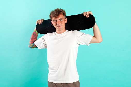 young cheerful tattooed guy in white t-shirt holding skateboard and smiling on blue isolated background, portrait of hipster skater