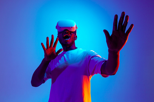 shocked african american male gamer in virtual reality glasses touches hands in neon lighting, frightened guy uses modern vr gadget and screams