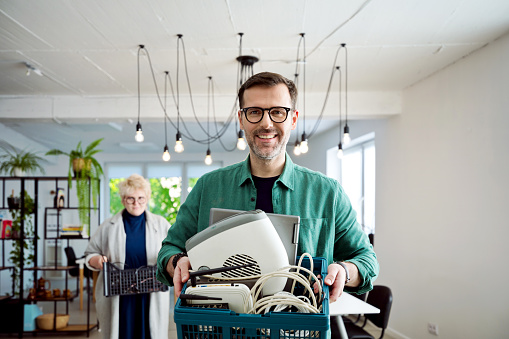 Mature man standing in the office and holding basket with e-waste, smiling at camera.