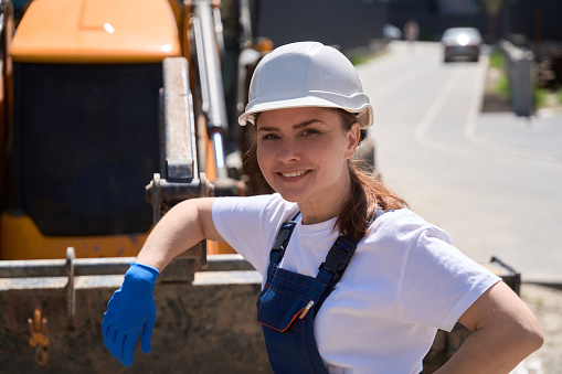 Smiling woman bulldozer operator leaning on blade, resting during hard working day at construction site, conducting in landscaping