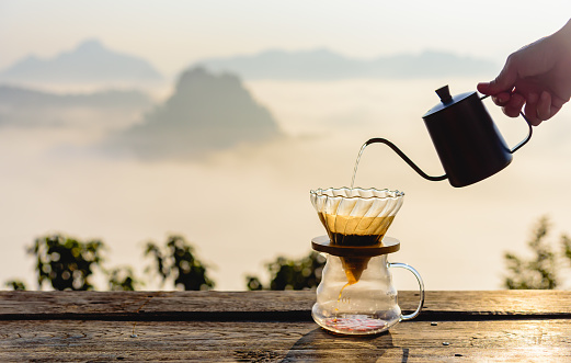 Hand pouring hot water on coffee ground with fillter, barista using drip coffee kit set with mountain and fog in sunrise background.