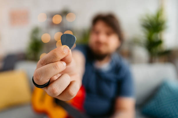 Overweight young man holding up a plectrum for a guitar at home in the living room.