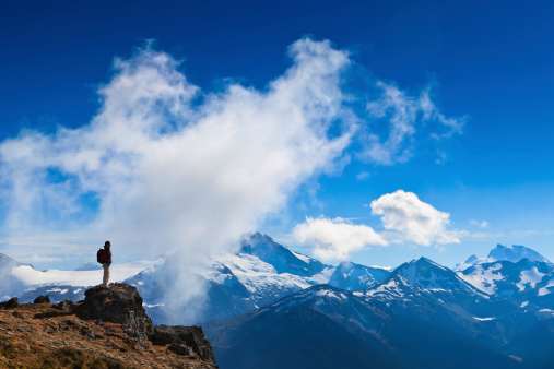 A woman is standing on a rock overlooking  the canadian rocky mountains when a cloud is going up the cliff just in front of her.