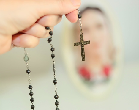 A hand holding old rosary beads against a defocused image of Our Lady