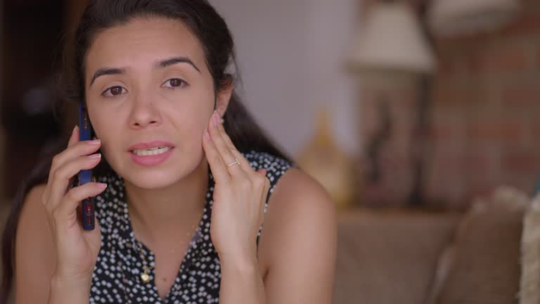 Sad Hispanic young woman feeling worried and stressed while talking on the phone at the livingroom. Bad news.