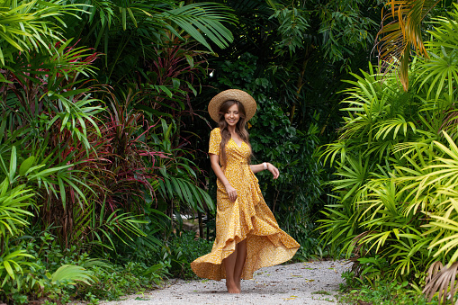 Female tourist girl with fashionable hat poses elegantly in lush green jungle. Young woman dancing charm and happiness in portrait of a tropical vacation.