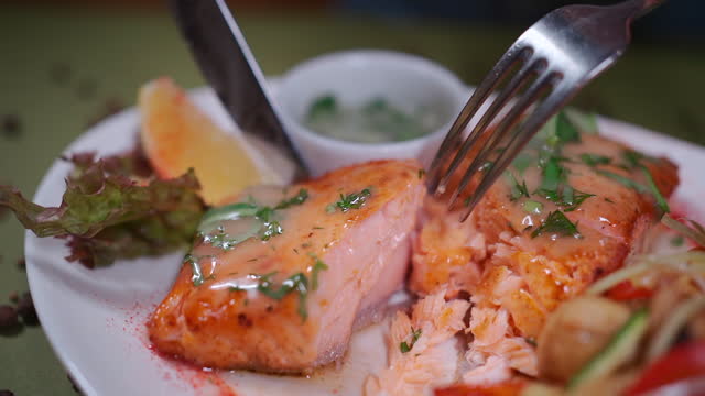 closeup of hand cutting roasted salmon steak on white plate with wok vegetable