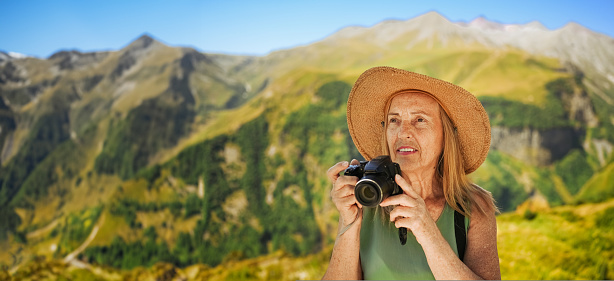 Portrait of a smiling happy elderly woman tourist traveling with a camera posing against the backdrop of mountains. Old elegant lady in straw hat on grass at countryside. Active retired people concept