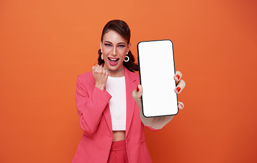 Happy celebration business woman wearing pink suit showing blank screen smart phone in hand isolated on orange background.