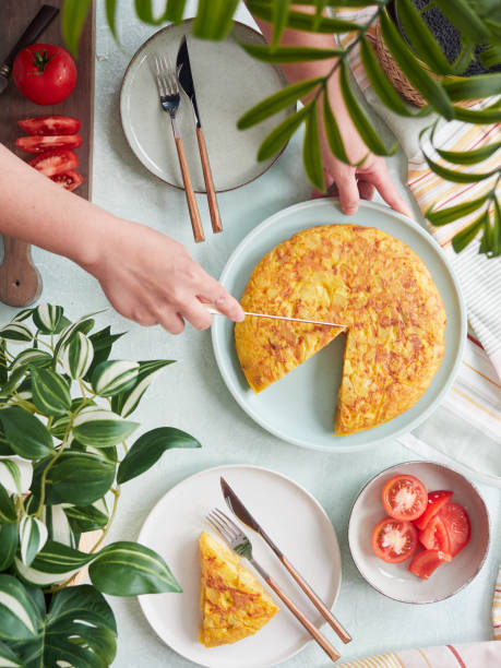 draufsicht auf eine person am tisch, die dreieckige portionen von typisch spanischem kartoffelomelett auf tellern mit tomatensalat serviert. - spanisches omelett stock-fotos und bilder