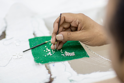 Detail closeup of a handicraft tailors hand embroidering small beads and crystals on tulle fabric to make a dress.
