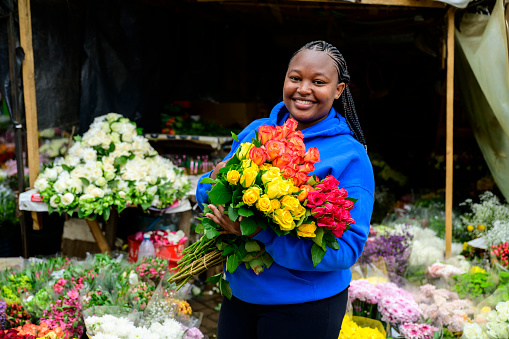 Portrait of mid 20s African woman smiling at camera while holding bouquets of red, yellow, and orange roses for sale in open-air market stall.
