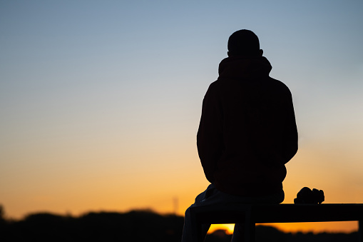 Silhouette of a man sitting on a bench against the backdrop of sunset. Copy space, headphones on the bench.