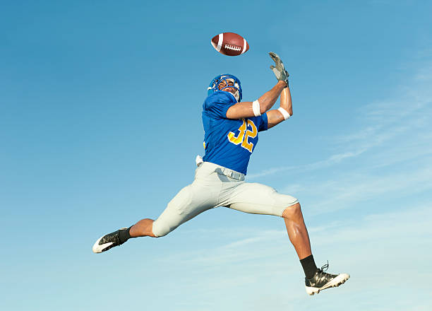 Mid-air shot of a player in blue catching a football Football player catches ball in midair. wide receiver athlete stock pictures, royalty-free photos & images