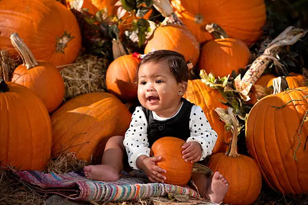 Happy baby girl sitting up in front of a pile of pumpkins holding a pumpkin.