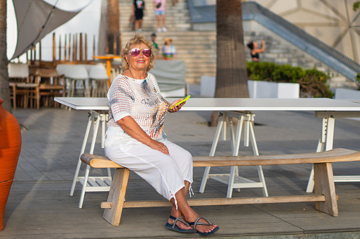 Smiling white haired senior woman reading messages on smart phone outdoor on a sunny day near the beach. Attractive elderly lady holding cellphone enjoying free time and vacation