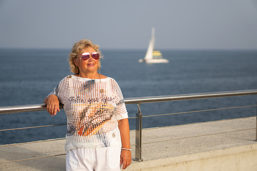 Smiling elderly woman with sunglasses walks along the beach near the sea on a sunny day. Active recreation, travel and freedom
