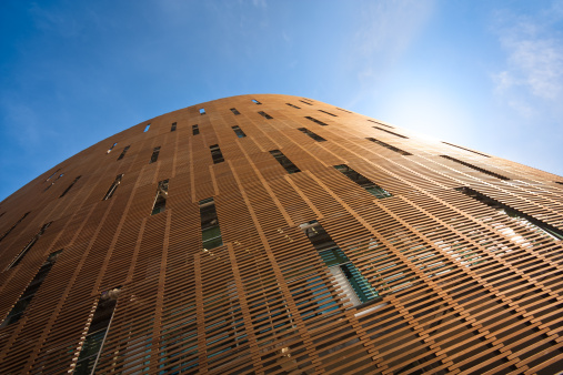 A modern building. It is partially rounded and has an external, grid like pattern which seems made of wood. Shot from the ground up against a bright blue sky.