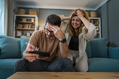 man adult caucasian male sit on sofa bed at home play video games on smartphone hold mobile phone while his wife or girlfriend in background is disappointed