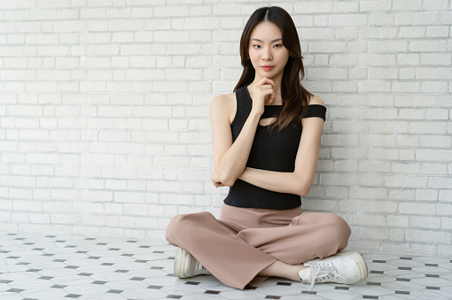 Portrait of beautiful young Asian woman wearing off shoulder top sitting on floor and looking at camera on white brick wall background. Millennials Korean or Japanese girl lifestyle.