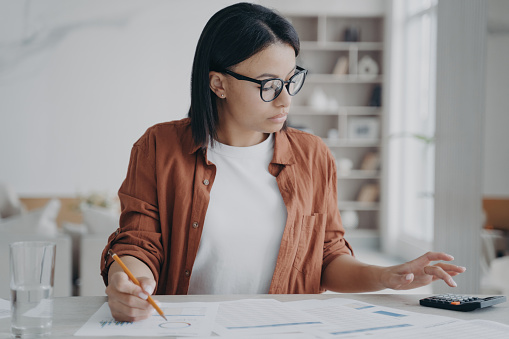 Focused businesswoman in glasses calculates expenses, plans company budget with a calculator. Dedicated female accountant freelancer works on financial reports. Effective financial management.