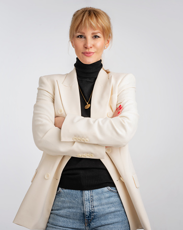 An attractive blond haired woman wearing white blazer and black sweater. Smiling female standing with arms crossed. She is against white background. Copy space.