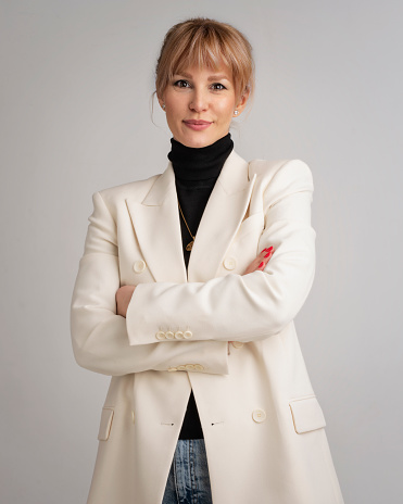 An attractive blond haired woman wearing white blazer and black sweater. Smiling female standing with arms crossed. She is against grey background. Copy space.