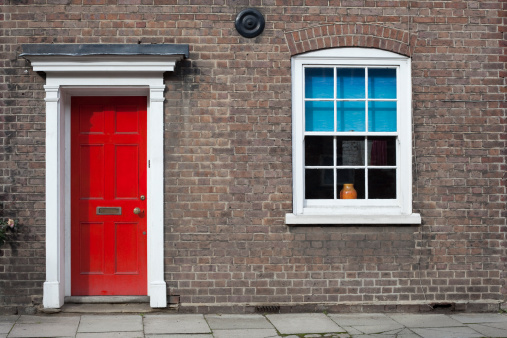 A red door, a window with blue blinds, and  white surrounds on a brick town house in a British village.