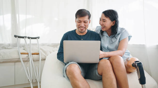 istock Happy asian woman in leg prosthesis and her husband sitting on comfortable sofa while typing on laptop for working online or chatting on social media at home. Leg prosthetic equipment 1537486688