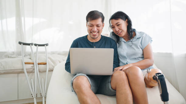 istock Happy asian woman in leg prosthesis and her husband sitting on comfortable sofa while typing on laptop for working online or chatting on social media at home. Leg prosthetic equipment 1537487202