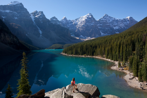 Female backpacker standing on boulder admiring majestic view of Moraine Lake and Canadian Rockies.