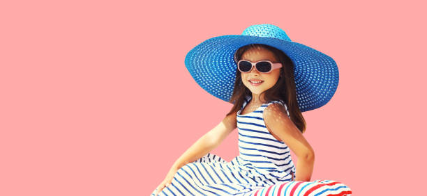 Summer vacation, beautiful little girl child in straw hat, striped dress sitting on pillow on pink background stock photo