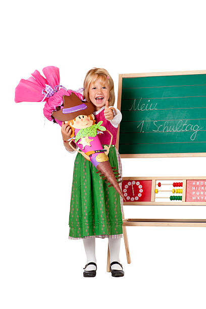 Girl on first school day stands beside chalk board and shows thumb. Isolated on white background.