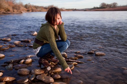 Cute teenage girl, crouching on river bank, dreamily lets a water deroplet fall from her finger